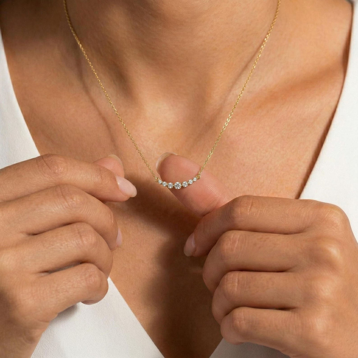 a close-up of a model wearing a delicate gold chain necklace with a small curved row of clear gemstones on the pendant, adjusting it with both hands against a white top.
