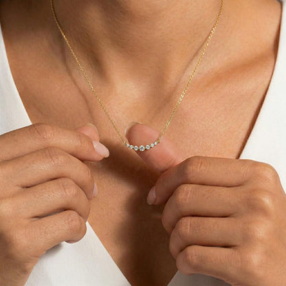 A close-up of a model wearing a delicate gold chain necklace with a small curved row of clear gemstones on the pendant, adjusting it with both hands against a white top.
