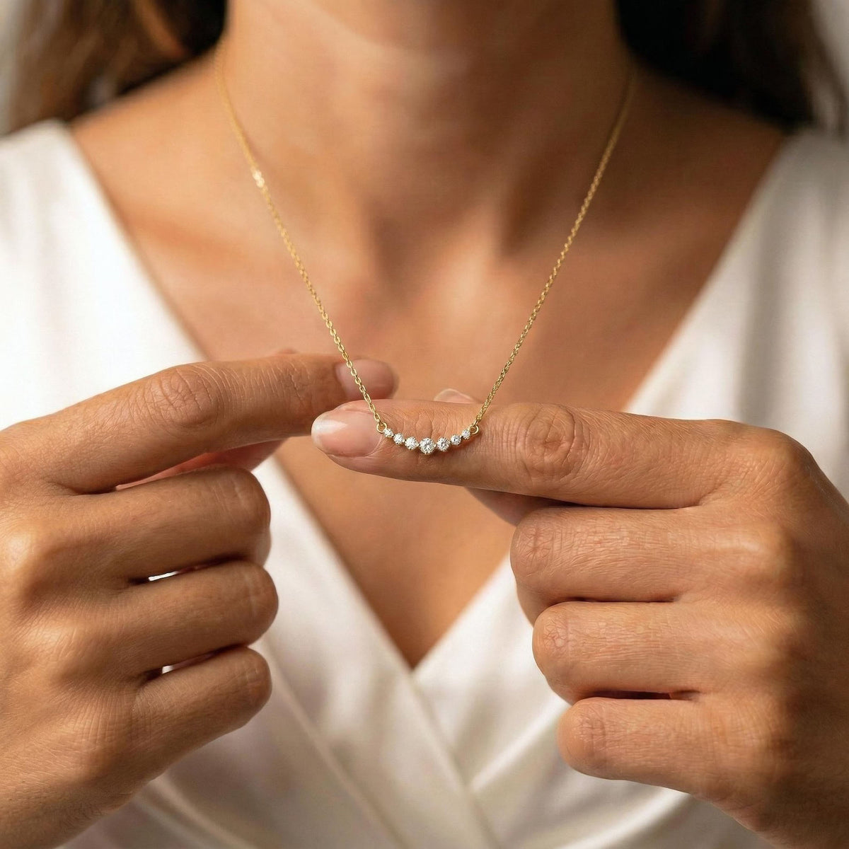 A close-up lifestyle shot of a model wearing a delicate gold chain necklace with a small curved row of clear gemstones on the pendant, held between her fingers.