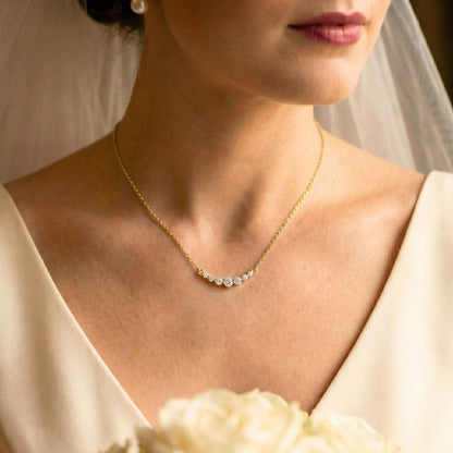 A close-up of a bride wearing a delicate gold necklace with a curved row of clear stones at the center of the chest. The image features a soft-focus background with a white bridal veil and cream-colored wedding dress.