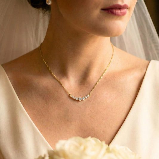 A close-up of a bride wearing a delicate gold necklace with a curved row of clear stones at the center of the chest. The image features a soft-focus background with a white bridal veil and cream-colored wedding dress.