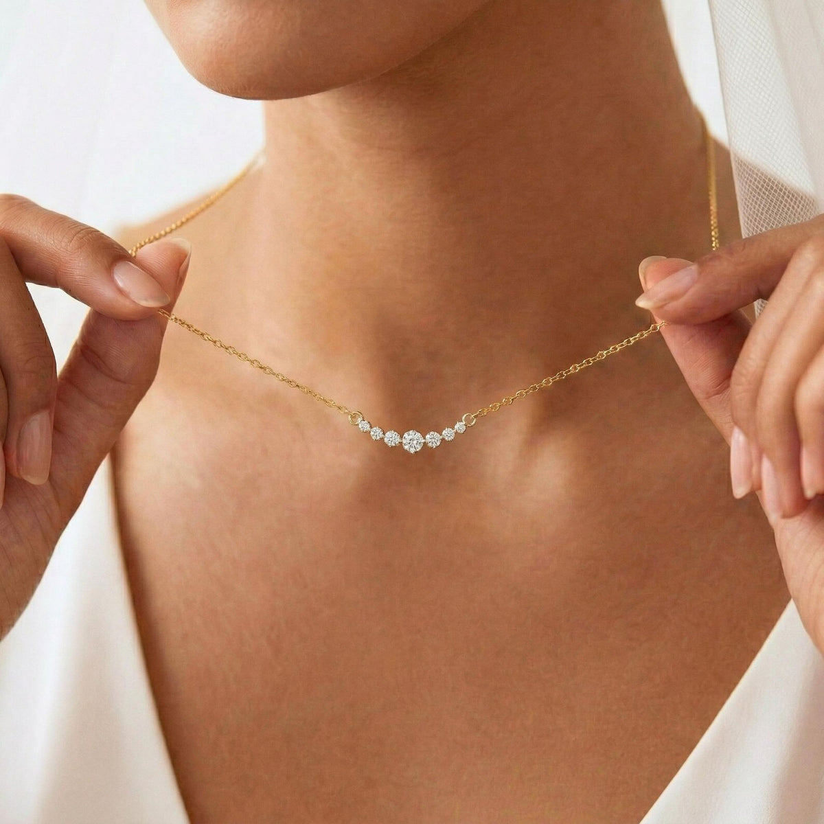 A close-up of a model wearing a delicate gold chain necklace with a vertical drop pendant featuring three clear round-cut gemstones, held gently by her hands against a soft white bridal veil.