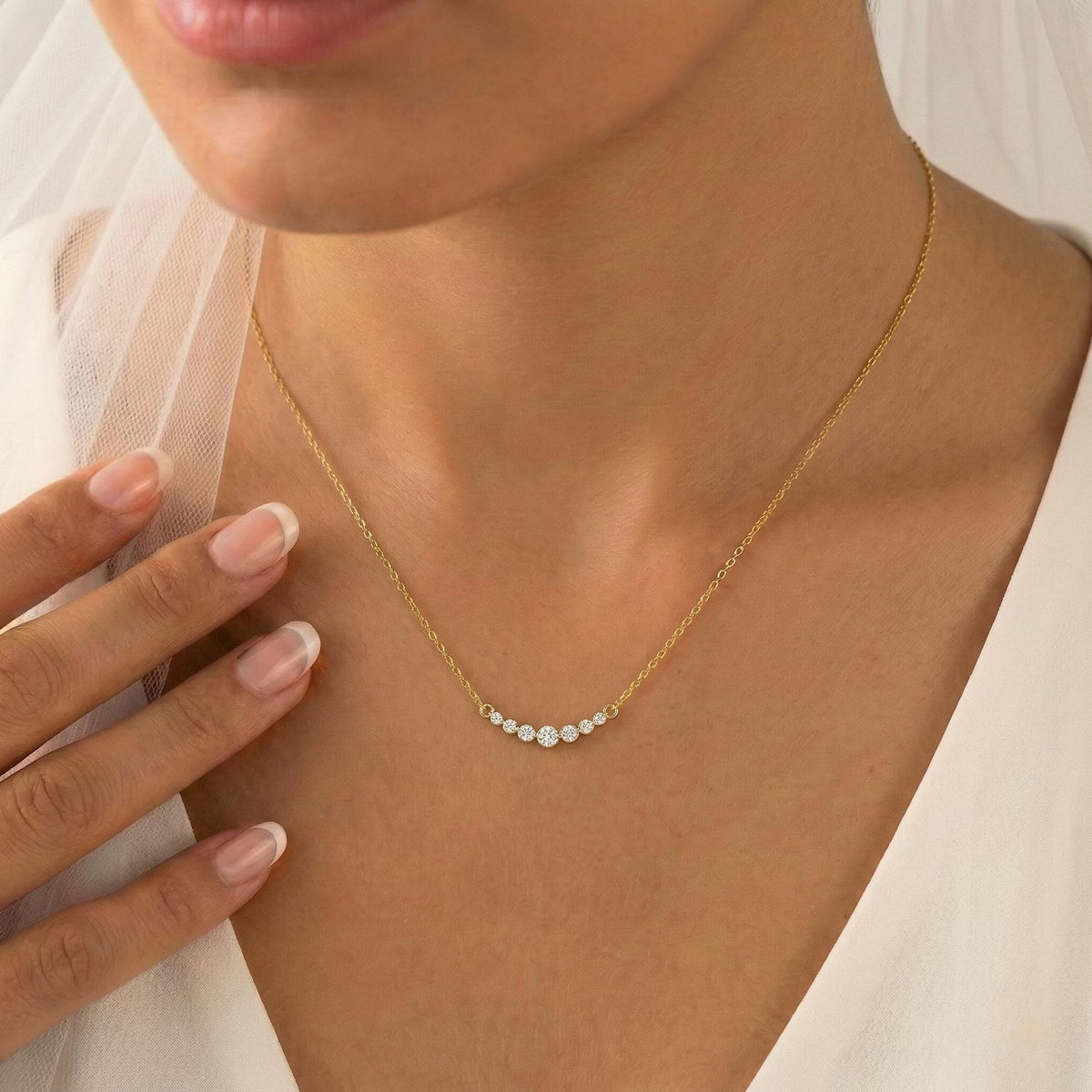 A delicate gold chain necklace with a curved row of small white pearl-like beads, worn on a model's neck. The model is wearing a white bridal veil and a white top, with her hand resting near her collarbone.
