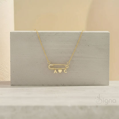 Signa™ Custom Gold Safety Pin Necklace featuring pave diamond accents on the pin head, two personalized initials, and a dangling heart charm. A dainty personalized birthday gift for her shown on a model.