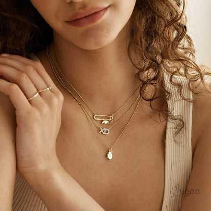 Signa™ Custom Gold Safety Pin Necklace featuring pave diamond accents on the pin head, two personalized initials, and a dangling heart charm. A dainty personalized birthday gift for her shown on a model.