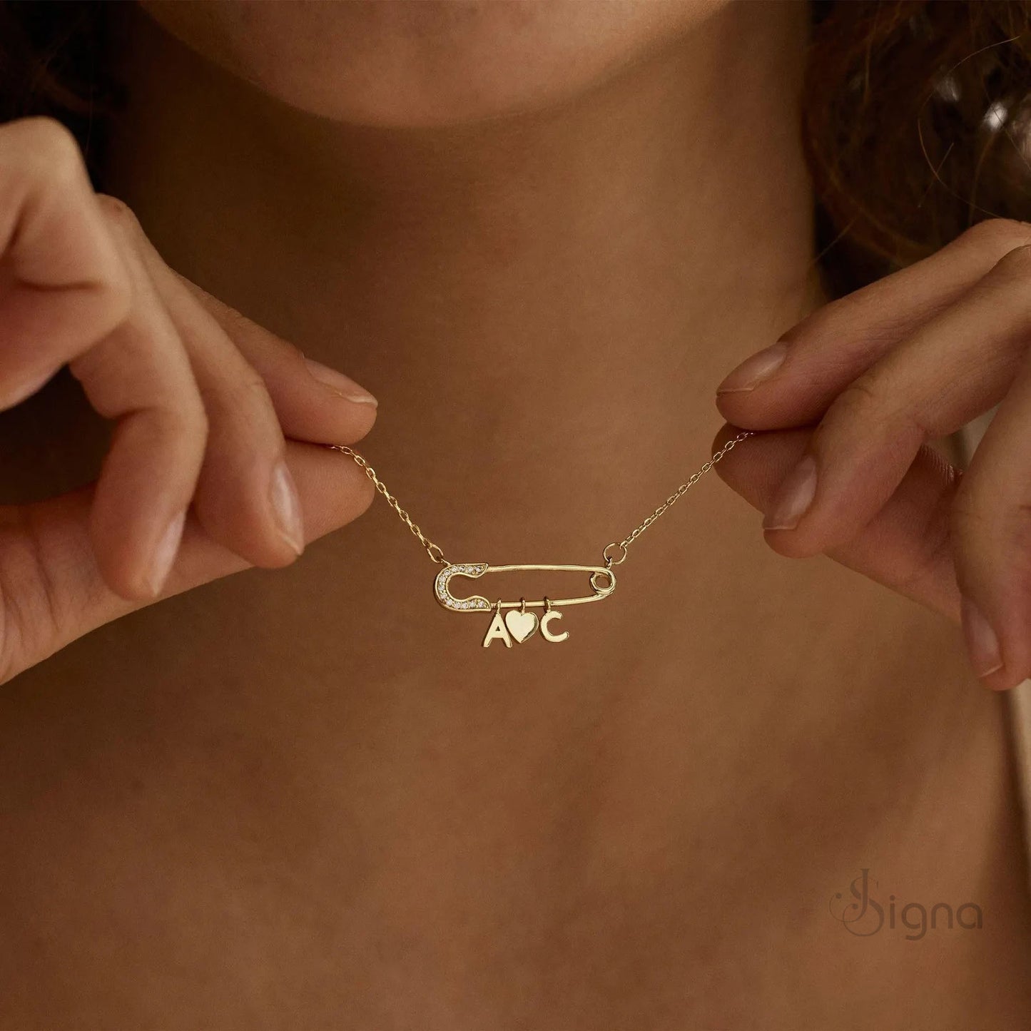signa™ custom gold safety pin necklace featuring pave diamond accents on the pin head, two personalized initials, and a dangling heart charm. a dainty personalized birthday gift for her shown on a model.