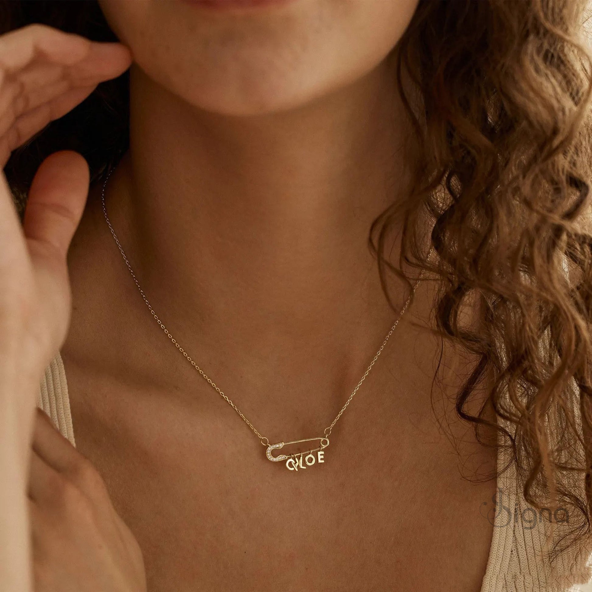 Signa™ Custom Gold Safety Pin Necklace featuring pave diamond accents on the pin head, two personalized initials, and a dangling heart charm. A dainty personalized birthday gift for her shown on a model.