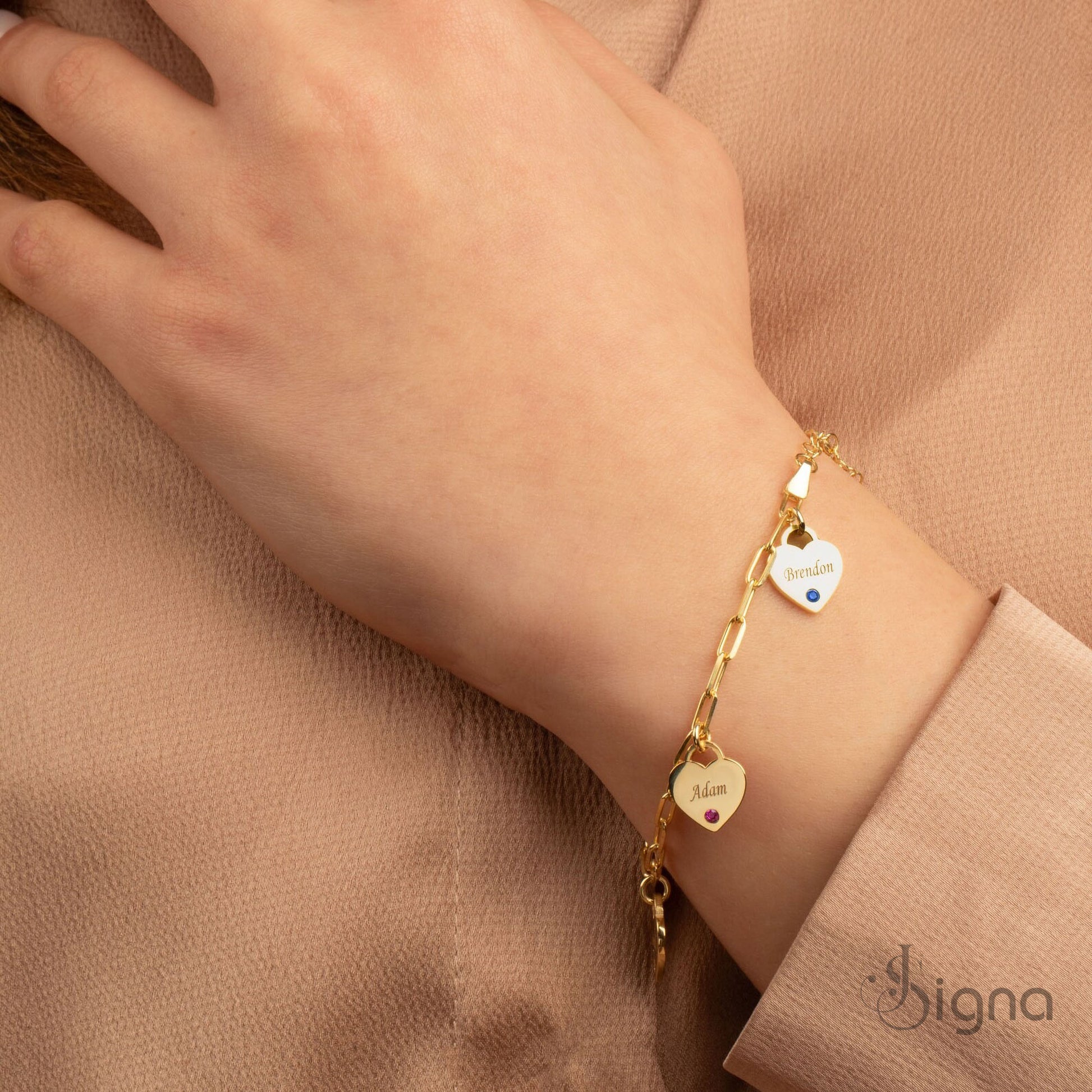 Gold bracelet with heart-shaped charms on a person's wrist against a beige background, featuring the brand 'igna'.